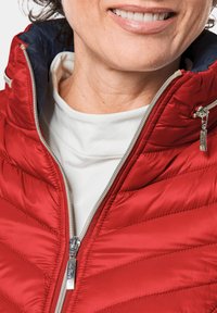 Doudoune matelassée rouge avec un col montant, une fermeture éclair argentée et une doublure bleu marine. Portée sur une chemise blanche, mettant en valeur sa texture légère.