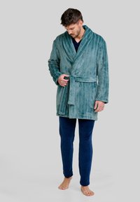 Bata de baño de forro polar a rayas en color azul verdoso con cierre de lazo, que presenta un cuello ancho y mangas largas, llevada sobre pantalones de terciopelo azul marino. Modelo descalzo.