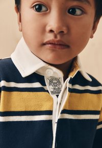 Polo avec un col blanc, présentant des rayures bleu marine, jaunes et blanches, fabriqué en tissu doux, avec une étiquette cousue visible au niveau de l'encolure.