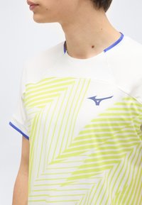 Νεαρό άτομο που φορά αθλητική μπλούζα με γεωμετρικό μοτίβο σε λευκό και λαχανί, με μπλε περιμετρικά και λογότυπο στο στήθος, κοιτάζοντας προς τα αριστερά.