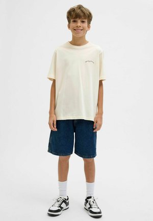 Helles beige Baumwoll-T-Shirt mit kurzen Ärmeln und einem kleinen bedruckten Design auf der Brust, kombiniert mit blauen Jeansshorts und schwarz-weißen Sneakers.