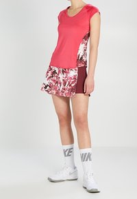 Mujer vestida con una camiseta deportiva rosa, una falda de estampado floral con pantalones cortos burdeos, calcetines blancos de Nike y zapatillas deportivas blancas, de pie sobre un fondo liso.
