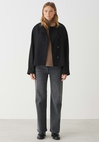 Giacca nera corta con ampio colletto, due bottoni e tasche laterali, indossata sopra un pullover beige e abbinata a jeans neri a gamba dritta.