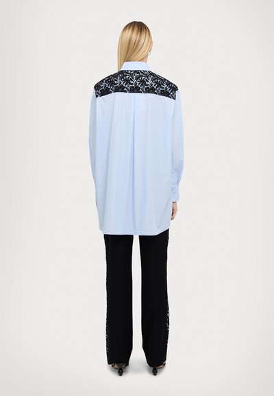 Camicia a maniche lunghe azzurro chiaro con yoke posteriore in pizzo nero, colletto button-down e vestibilità rilassata. Abbinata a pantaloni neri.