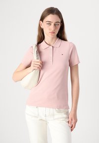 Ανοιχτό ροζ πόλο μπλούζα με κλασικό γιακά και κουμπιά. Συνδυασμένη με άσπρο παντελόνι και κρέμα τσάντα ώμου. Διακριτή λεπτομέρεια λογότυπου ορατή.