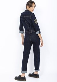 Dżinsowy kombinezon o dopasowanym kroju, w ciemnoniebieskim kolorze, z podwiniętymi mankietami i naszywką na lewym rękawie. Noszony z czarnymi sznurowanymi butami.