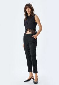 Zwart mouwloos topje met een doorschijnende onderoverlay, gecombineerd met op maat gemaakte zwarte broeken en glanzende zwarte slingback hakken.