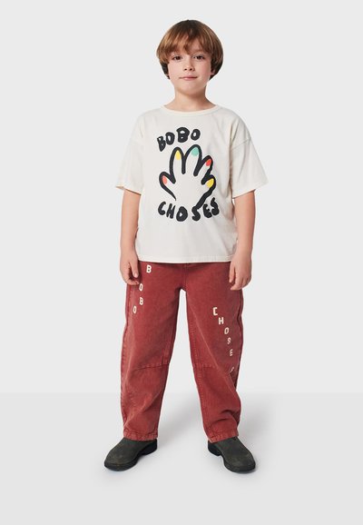 Vit bomullströja med färggrafik av händer och text. Lätta röda kordbyxor med "BOBO" och "CHOSES" tryckt på sidorna. Svarta skor.