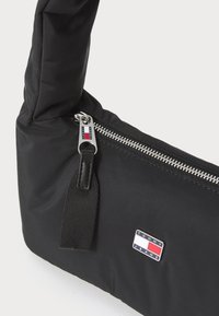 Crna Tommy Jeans torba s srebrnim zatvaračem, crnom tekstilnom povlačnom trakom i malim logotipom u crvenoj, bijeloj i plavoj boji.