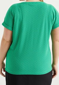 Grön kortärmad topp med en texturerad fiskbensmönster och rundad nederkant. Tyget är mjukt med en något lös passform.