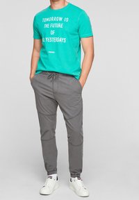 Man i en teal T-shirt med vit text, grå dragkedjebyxor och vita sneakers, står med händerna i fickorna mot en grå bakgrund.