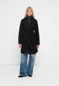 Cappotto nero con ampio colletto, vita cinturata e maniche lunghe, indossato sopra un giubbotto imbottito nero. Abbinato a jeans azzurri svasati.