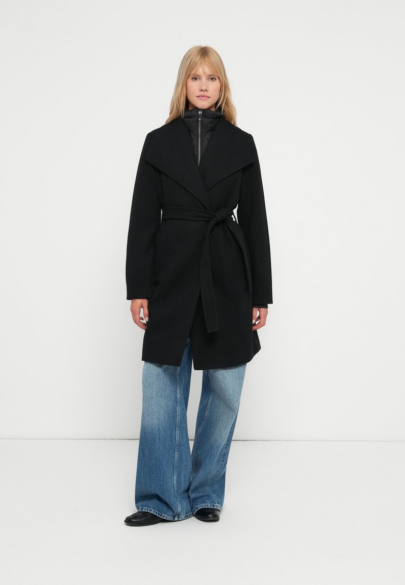 Cappotto nero con ampio colletto, vita cinturata e maniche lunghe, indossato sopra un giubbotto imbottito nero. Abbinato a jeans azzurri svasati.