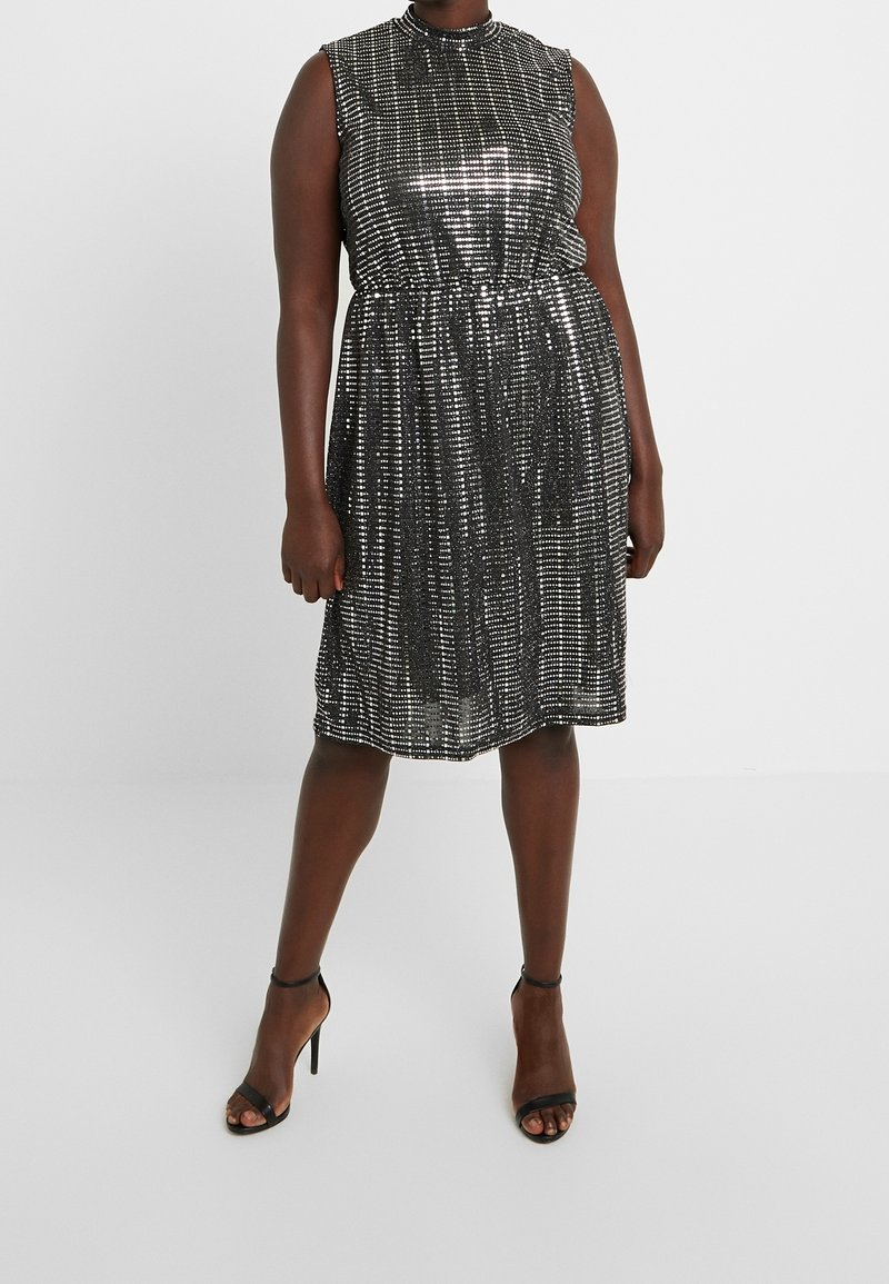 Femme portant une robe sans manches noire et argentée à sequins longueur genou, avec des talons hauts à bride à la cheville, debout devant un fond uni.