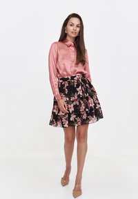 Różowa satynowa bluzka z kołnierzykiem zapinanym na guziki, zestawiona z czarną spódnicą z falbanami, ozdobioną kwiatowym nadrukiem w różu i zieleni. Przezroczyste obcasy.