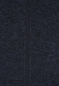 Marineblåt strikket sweaterstof med et tekstureret mønster. Midtercentreret lynlåslukning med mørkt lynlåsbånd, der giver et strømlinet udseende.