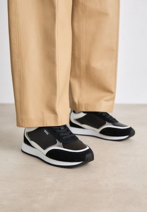 Osoba ubrana w beżowe spodnie i czarno-białe sneakersy BOSS, stojąca na jasnej podłodze.