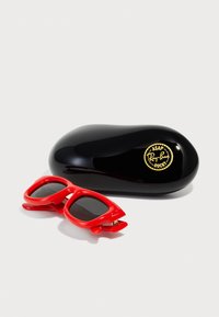 Røde solbriller med firkantede linser, med en unik form og design, liggende ved siden af et blankt sort etui med et guld logo akcent.