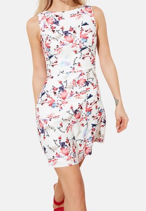 Frau trägt ein ärmelloses weißes Kleid mit roten, violetten und blauen Blumenmustern, kombiniert mit roten offenen Schuhen mit Absätzen.