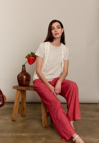 T-shirt gris clair avec dentelle, associé à un pantalon large rouge texturé présentant un motif ondulé. Fond beige et tabouret en bois.