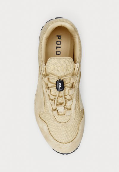 Beige színű sneaker, amely textúrázott felsőrészzel, velúr részletekkel, rugalmas fűzőrendszerrel és egy toggle-záróval, valamint fekete gumitalppal rendelkezik. "POLO" felirat a nyelvén.