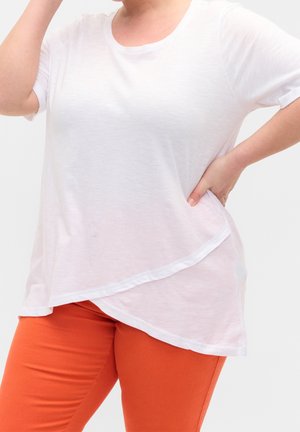 Osoba nosząca luźny biały top z krótkim rękawem o asymetrycznym kroju i jaskrawo pomarańczowe dopasowane spodnie, z ręką na biodrze.