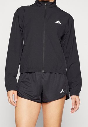 Zwarte sportjas met hoge kraag, volledige ritssluiting en wit logo. Gekombineerd met zwarte shorts met een split aan de zijkant en logo-detail.