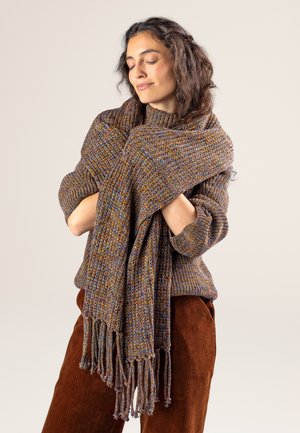 Multicolored knitted scarf with textured pattern and fringe edges, wrapped around a sweater-clad figure. Rich hues of brown, gold, and blue.