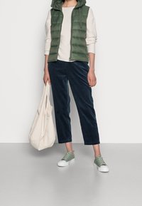 Groene gewatteerde bodywarmer over een crèmekleurige trui, gecombineerd met donkerbruine ribfluwelen broek en mintgroene sneakers. Beige draagtas in de hand.