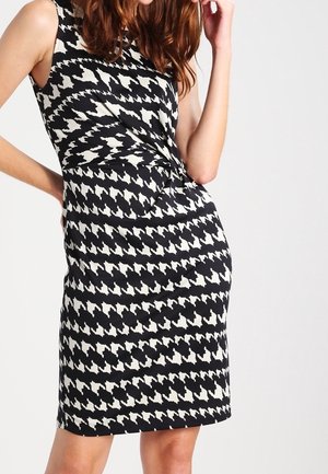 Robe en pied-de-poule noir et blanc avec une silhouette ajustée, sans manches, et un détail torsadé à la taille. Texture lisse.