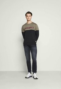 Um jovem está de pé, vestindo um sweatshirt colorido com o texto "ORIGINALS", jeans escuros, meias pretas e ténis brancos, contra um fundo simples.