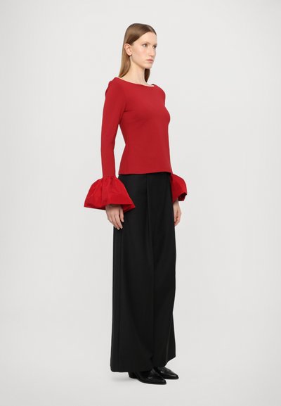 Red long-sleeve top with oversized ruffled cuffs, paired with black wide-leg trousers and black pointed-toe shoes. Smooth fabric, fitted design.