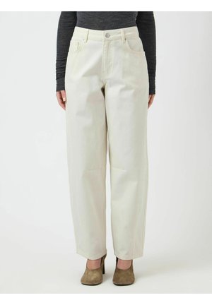 Vrouw draagt een off-white, hooggetailleerde broek met wijde pijpen, gecombineerd met bruine suède hakken en een donker shirt met lange mouwen, staand tegen een effen achtergrond.