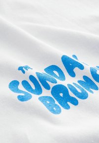 Fehér pamut póló, texturált kék nyomtatott grafikával, amelyen a "THE SUNDAY BRUNCH" felirat szerepel játékos, kerek betűtípusban.