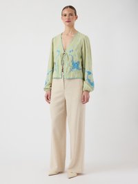 Kvinne iført lys grønn langermet bluse med blå broderi og beige vide bukser, stående mot en ensfarget hvit bakgrunn.