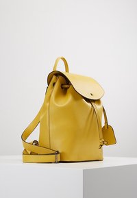 Sac à dos en cuir jaune avec une texture lisse, une forme arrondie, un rabat pliant, une sangle réglable et une petite étiquette attachée.
