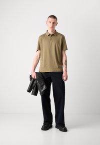 Polo khaki avec une patte de boutonnage à trois boutons, associé à un jean bleu foncé. Le mannequin tient une veste en cuir noir, se tenant dans un cadre neutre.