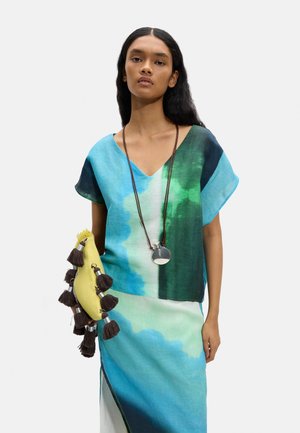 Kobieta ubrana w bluzkę z dekoltem w serek o gradientowym kolorze niebiesko-zielonym oraz spódnicę, trzymająca żółtą kopertówkę z brązowymi frędzlami i srebrny naszyjnik z zawieszką.