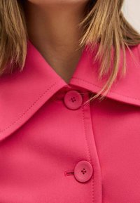Blusa de color rosa con grandes botones redondos, textura suave y un diseño estructurado. Líneas limpias con un ligero brillo.