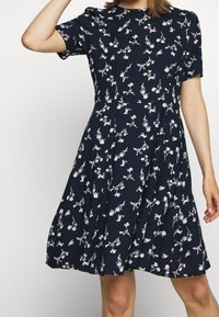 Kvinna iklädd en marinblå knälång klänning med korta ärmar och ett vitt blommönster, poserande med en arm upplyft.