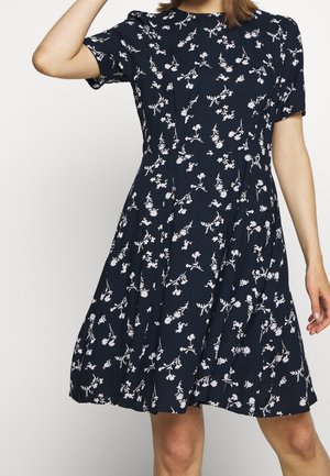 Frau in einem marineblauen knielangen Kleid mit kurzen Ärmeln und einem weißen Blumenmuster, die mit einem erhobenen Arm posiert.
