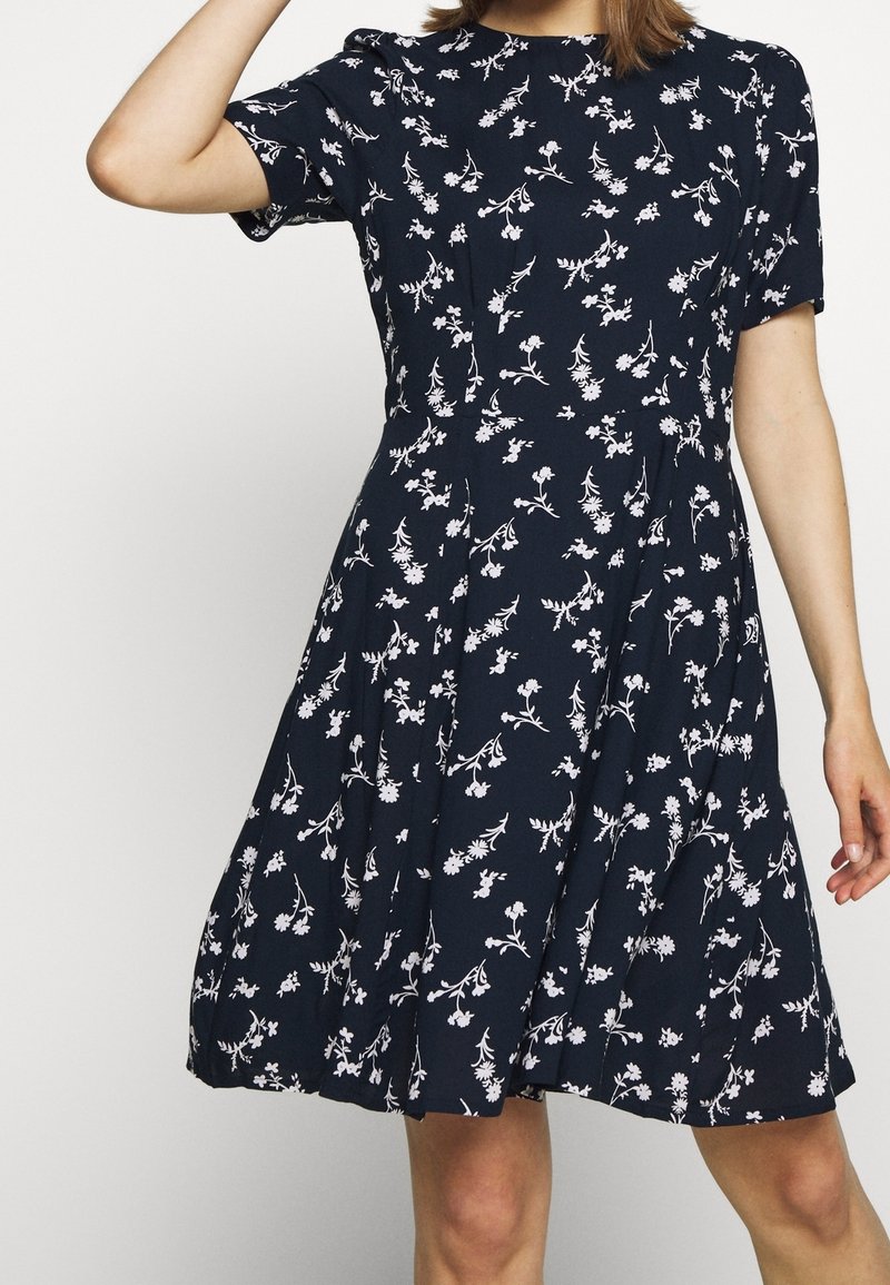 Kvinna iklädd en marinblå knälång klänning med korta ärmar och ett vitt blommönster, poserande med en arm upplyft.