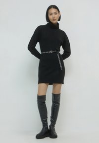 Schwarzes gestricktes Sweaterkleid mit hohem Kragen, ausgestattet mit einem silbernen Gürtel mit Ösen und kniehohen schwarzen glänzenden Stiefeln.