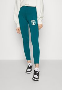 Turfärgad leggings med hög midja och vit Nike-logotypdetalj på vänster lår, i kombination med svart och vita träningsskor.