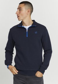 Muškarac s kratkom kosom nosi tamnoplavu duksericu sa četvrtastim zipom, plavim patentnim zatvaračem i logotipom, gleda ulijevo s rukama u džepovima traperica.