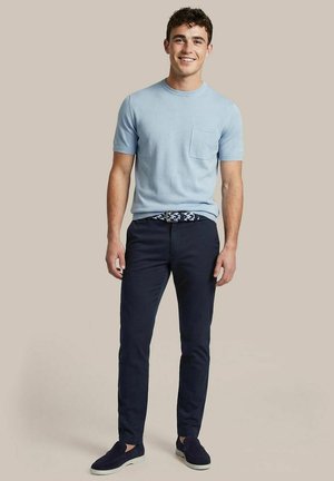 Jonge man die staat en lacht, gekleed in een lichtblauw overhemd met korte mouwen en een zakje, donkerblauwe broek, patroonriem en donkere instapschoenen.