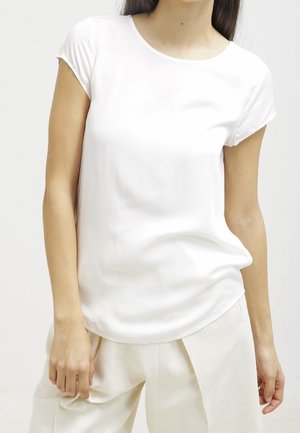 Vrouw draagt een effen wit t-shirt met korte mouwen en losse witte broek, staat met één hand ontspannen langs haar zijde.