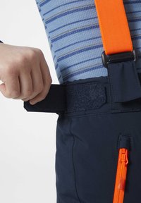 Los pantalones impermeables de color navy cuentan con una correa negra ajustable de Velcro y un bolsillo lateral con cremallera naranja. La capa interior presenta rayas azules.