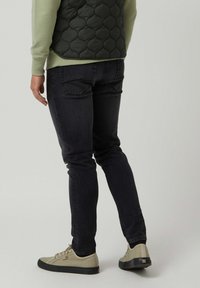 Persona con jeans ajustados negros, zapatillas beige con suelas negras, una camisa verde de manga larga y un chaleco negro acolchado.