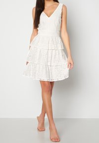 Abito bianco in pizzo con scollatura a V, design senza maniche e gonna a strati. Il tessuto è texturizzato e presenta un motivo floreale, con un punto vita aderente.